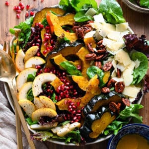 A large round platter filled with Thanksgiving Salad made with pears, pomegranate seeds, roasted acorn squash, candied pecans, Parmesan cheese and mixed greens. The toppings are arranged over the greens in rows.