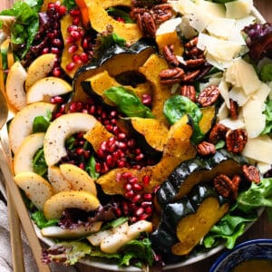 A large round platter filled with Thanksgiving Salad made with pears, pomegranate seeds, roasted acorn squash, candied pecans, Parmesan cheese and mixed greens. The toppings are arranged over the greens in rows.