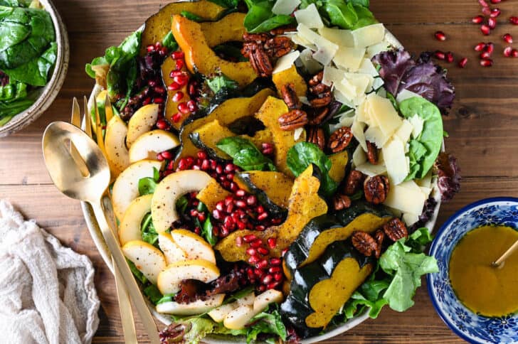 A large round platter filled with the best Thanksgiving Salad made with pears, pomegranate seeds, roasted acorn squash, candied pecans, Parmesan cheese and mixed greens. The toppings are arranged over the greens in rows.