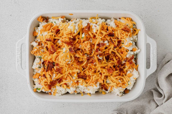 A white rectangle baking dish filled with a light colored casserole mixture topped with shredded cheese and bacon.