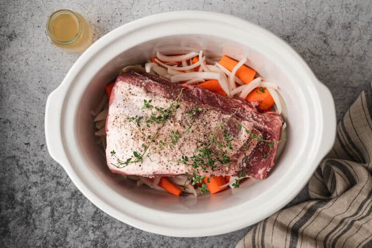 A white slow cooker insert with sliced carrots, onions and garlic with a pork roast on top inside.