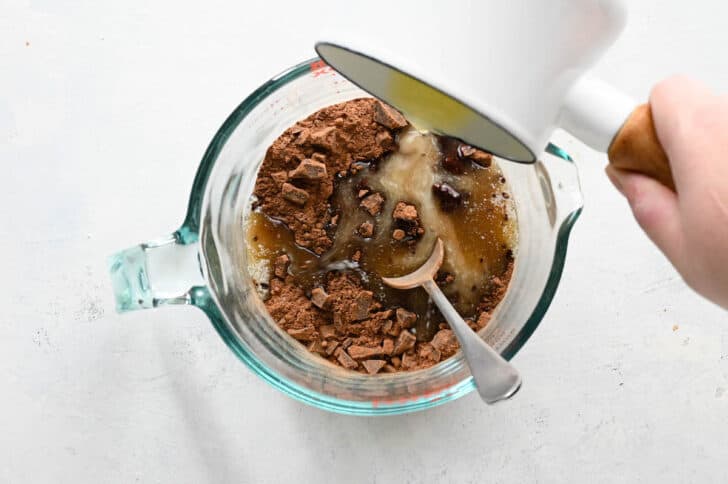 A hand pouring melted butter over a large glass measuring cup filled with cocoa powder and chopped chocolate.