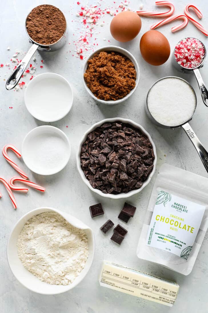 Ingredients laid out on a light surface, including flour, sugar, brown sugar, cocoa powder, eggs, chopped chocolate from a bag of Harvest Chocolate Haiti Snacking Chocolate, and candy canes.