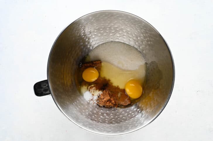 A bowl of a stand mixer will two eggs, sugar, brown sugar and salt inside the bowl.