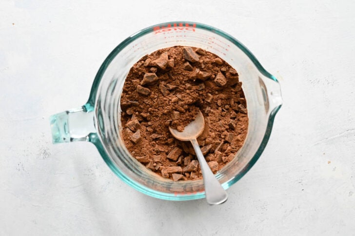 A large glass measuring cup with a spoon stirring together cocoa powder and chopped chocolate.