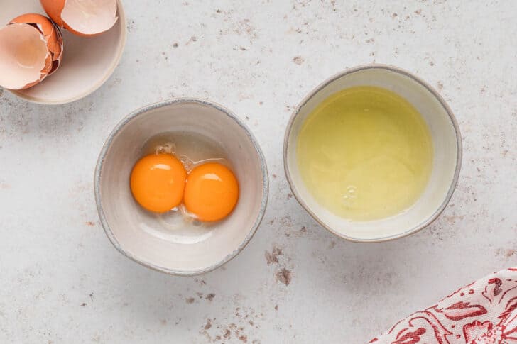 Two small white bowls. One is filled with two egg yolks, and the other has the egg whites in it.
