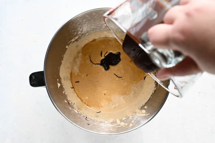 A hand pouring melted chocolate into a light brown batter mixture inside the bowl of a stand mixer.