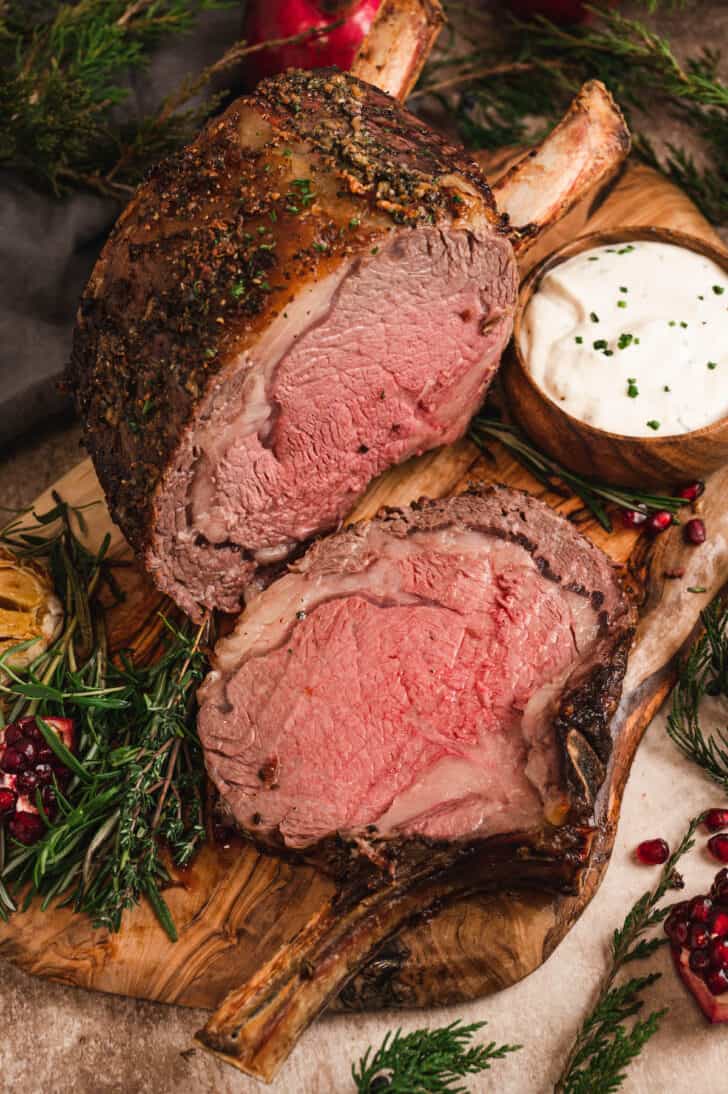 A medium-rare prime rib that has had one piece sliced off of it, on a wooden cutting board with a bowl of creamy horseradish sauce.
