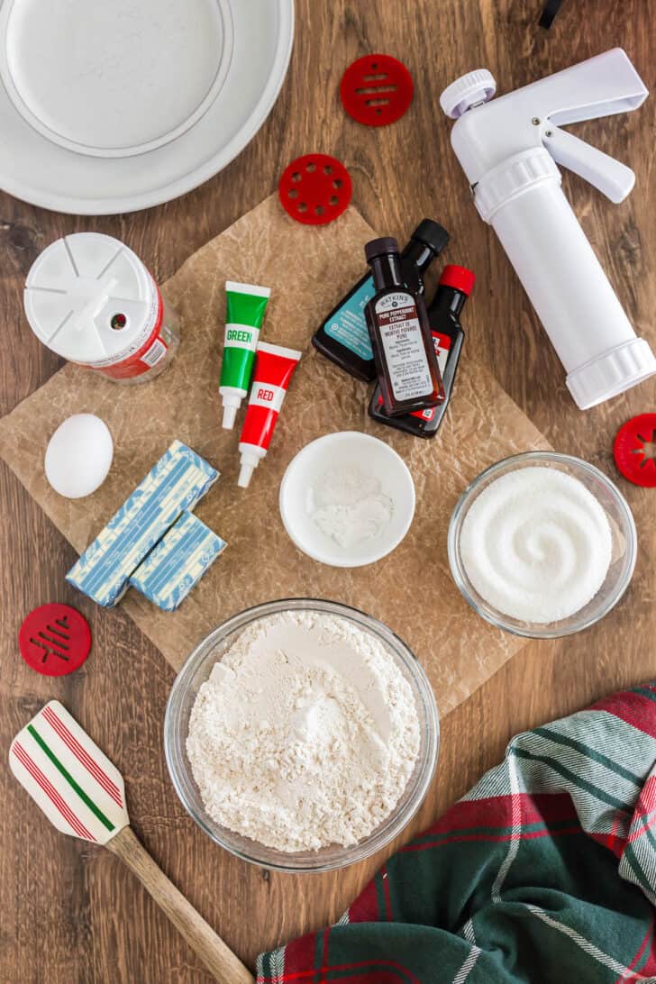 The ingredients needed for a spritz cookies recipe, including flour, sugar, food coloring, butter, and egg, along with a cookie press.
