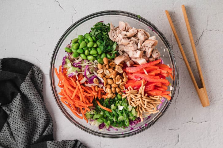 A glass bowl filled with chicken, red peppers, chow mein noodles, green onions, cilantro, carrots, cabbage, edamame and roasted peanuts.