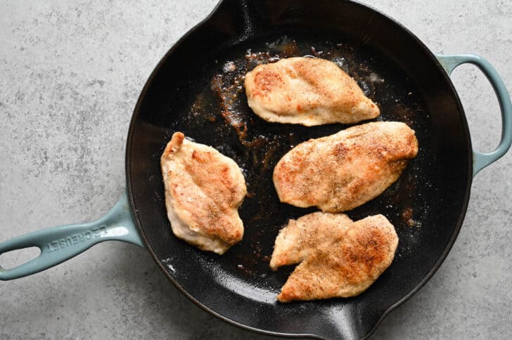 Four chicken breasts being cooked in a cast iron skillet.