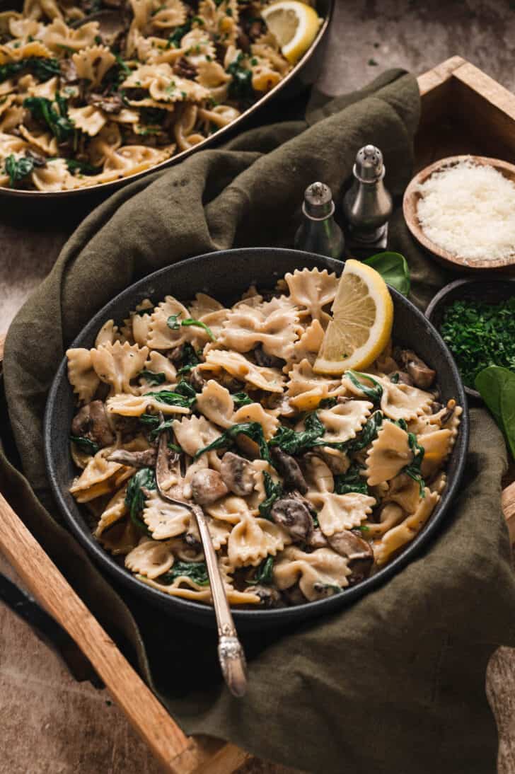 A dark bowl filled with a creamy mushroom pasta recipe, on a linen-covered tray with garnishes.