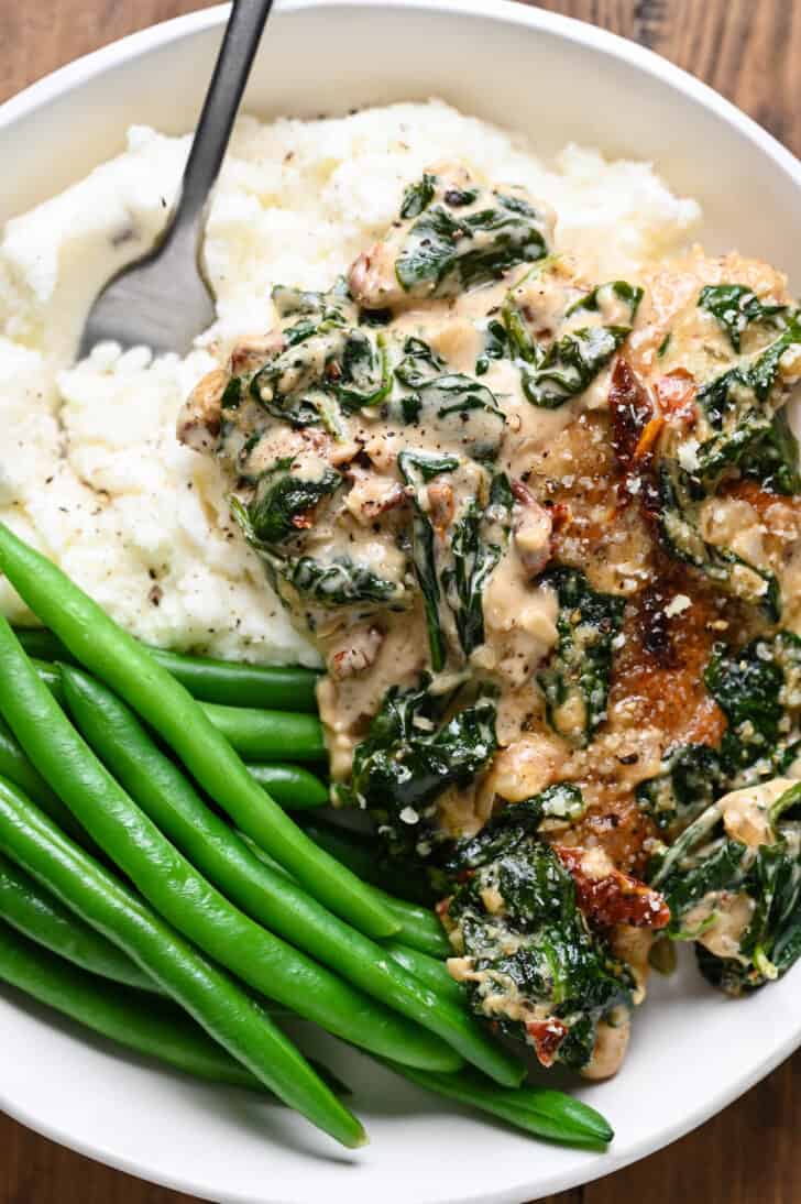 A white bowl filled with mashed potatoes, green beans and chicken with spinach cream sauce.