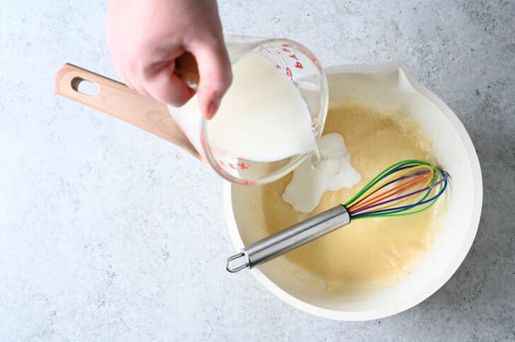 A white saucepan filled with roux, with a colorful whisk stirring it, with a hand pouring in milk.