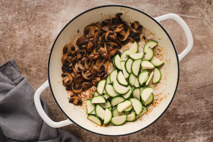 A white Dutch oven filled with cooked mushrooms and raw zucchini slices.