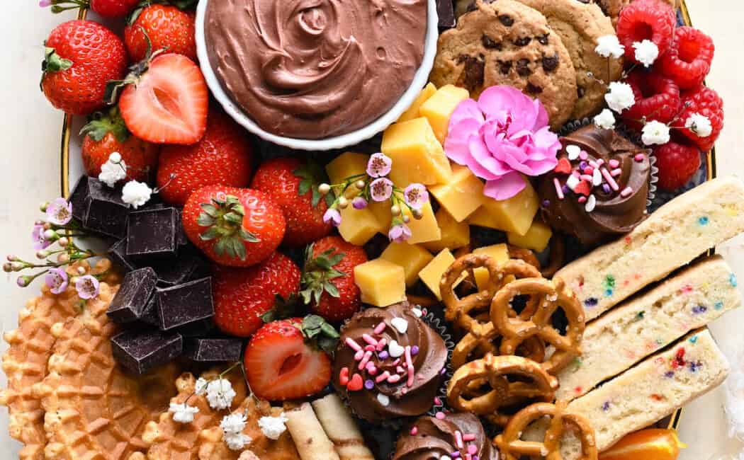 A dessert charcuterie board featuring chocolate dip, cookies, fruit, pretzels, cupcakes and flowers.