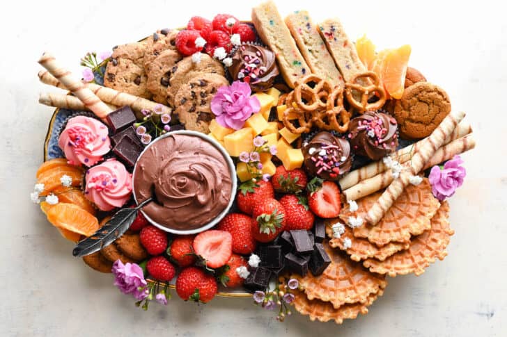 A charcuterie board dessert featuring chocolate dip, cookies, fruit, pretzels, cupcakes and flowers.