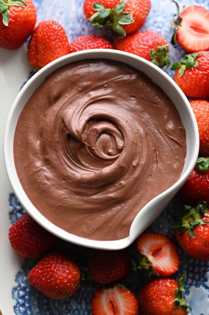 A white bowl filled with chocolate fruit dip, surrounded by strawberries.