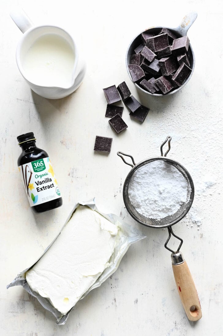Ingredients on a light surface, including chocolate chunks, milk, vanilla extract, powdered sugar and cream cheese.