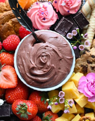A white bowl filled with chocolate dip, surrounded by fruit, cheese, cookies, chocolate and flowers.