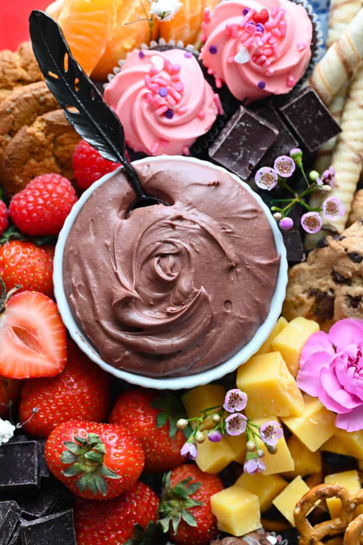 A white bowl filled with chocolate dip, surrounded by fruit, cheese, cookies, chocolate and flowers.