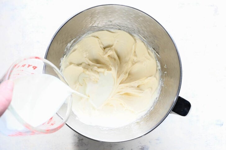 A stainless steel stand mixer bowl filled with a beaten cream cheese mixture, with a hand pouring milk in.