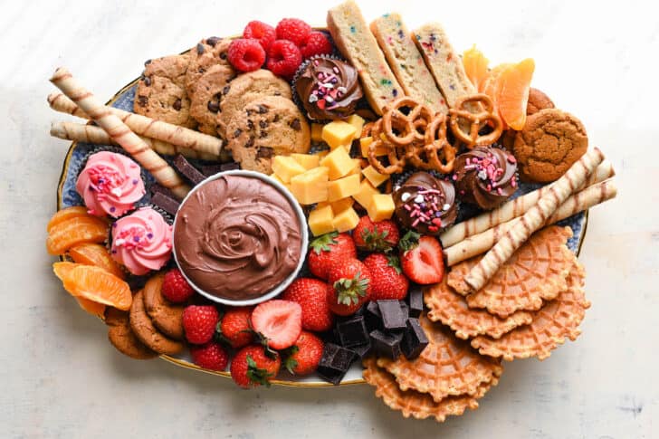 A dessert charcuterie board featuring chocolate dip, cookies, fruit, pretzels and mini cupcakes.