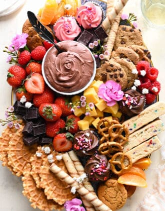 A dessert charcuterie board featuring chocolate dip, cookies, fruit, pretzels, cupcakes and flowers.