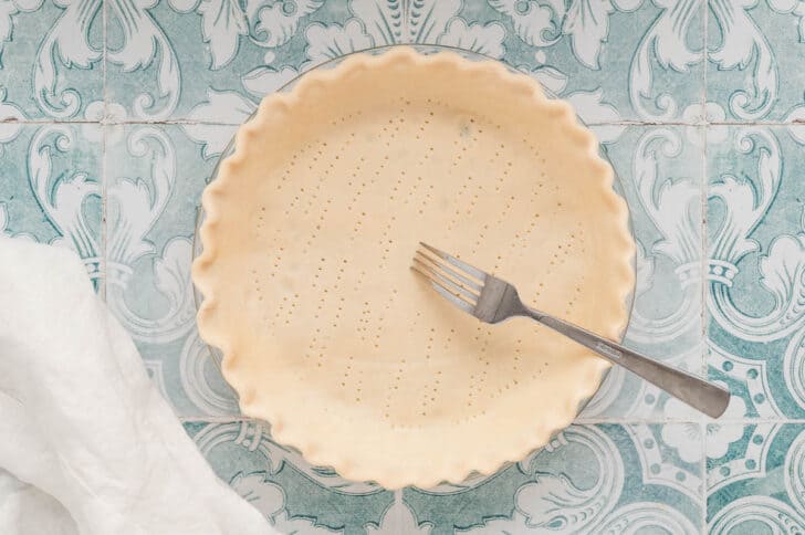 A pie plate filled with a fluted pie crust, with a fork making docking holes in the bottom