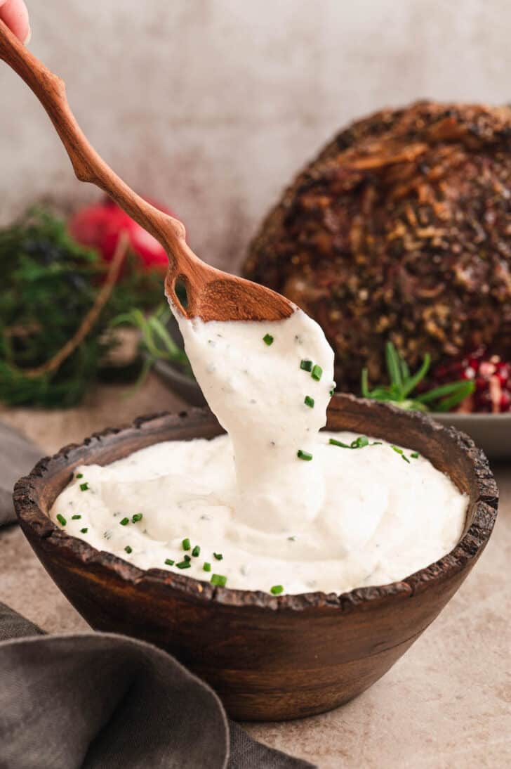 A rustic wooden bowl filled with horseradish cream sauce, with a wooden spoon lifting some from the bowl.