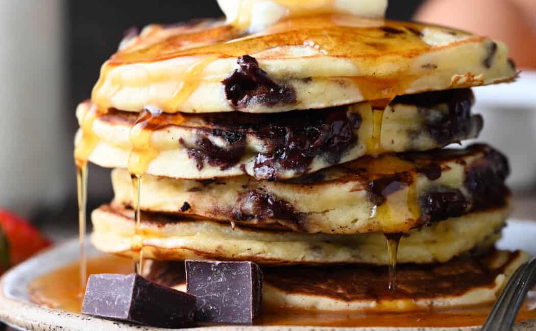 A stack of chocolate chip pancakes with a pat of butter on top and syrup being poured over them.