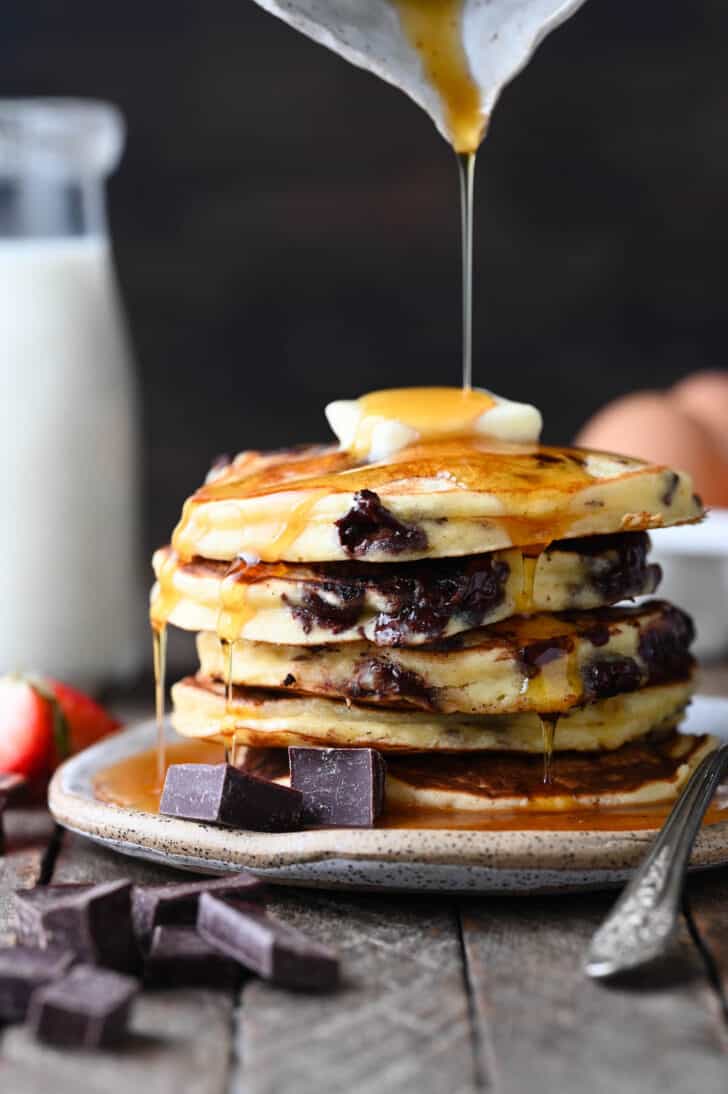 A stack of chocolate chip pancakes with a pat of butter on top and syrup being poured over them.