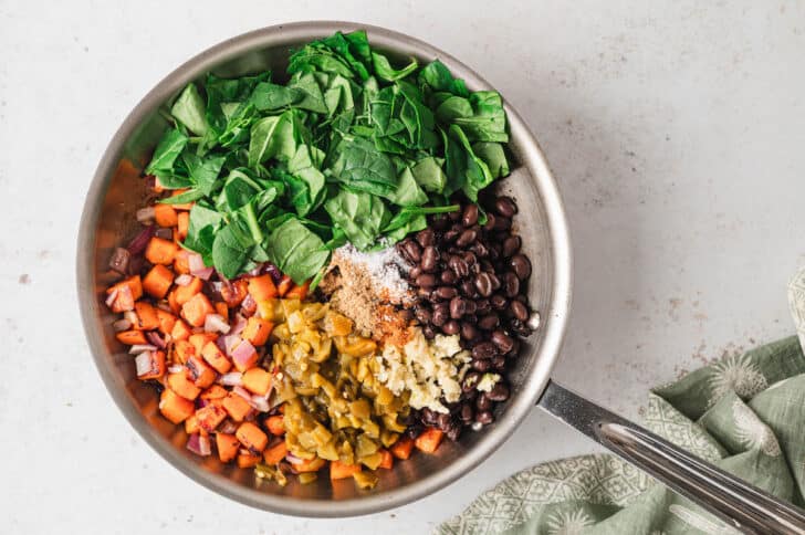 A stainless steel skillet filled with black beans, a sweet potato mixture, spinach, green chiles and spices.