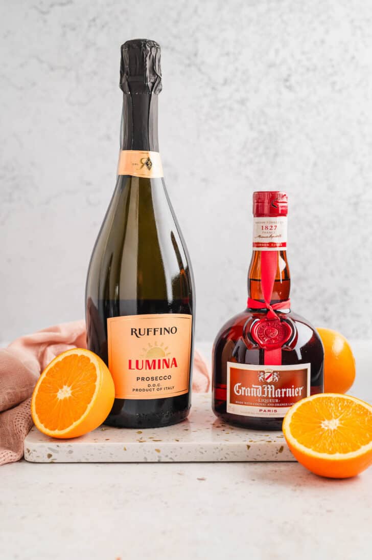 Ingredients needed for how to make mimosas, including bottles of prosecco and Grand Marnier, along with fresh orange halves.