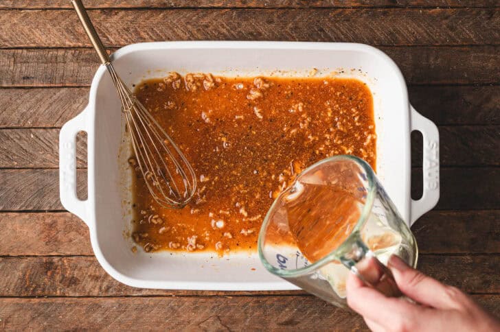 A hand pouring oil into a baking dish filled with an orange sauce.
