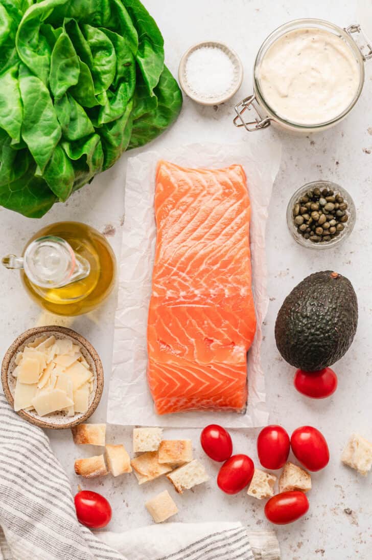 Ingredients on a light background, including raw salmon, avocado, tomatoes, bread cubes, Parmesan cheese, lettuce, capers, oil, salt and creamy salad dressing.