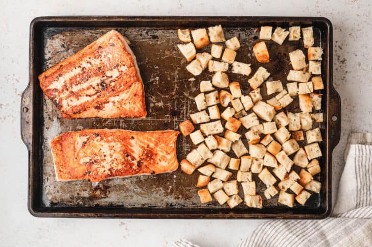 A rimmed baking pan with two salmon fillets on one half and toasted bread cubes on the other half.