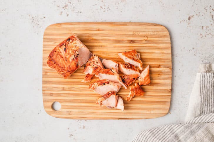 A wooden cutting board topped with a cooked salmon fillet being flaked into large pieces.