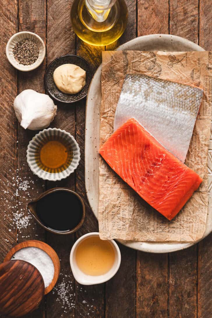 Ingredients on a wooden surface, including raw fish, garlic, oil, spices and condiments.