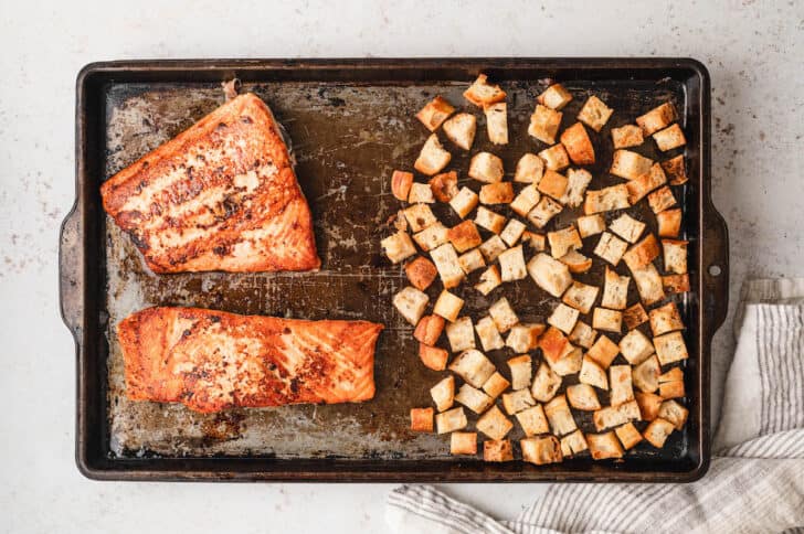 A rimmed baking pan with two salmon fillets on one half and toasted bread cubes on the other half.