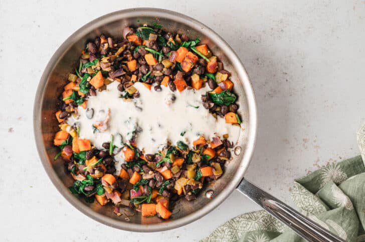 A stainless steel skillet filled with a sweet potato and black bean mixture, topped with a spoonful of creamy sauce.