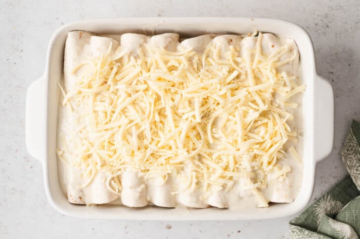 A rectangle white baking dish filled with rolled up flour tortillas topped with creamy sauce and shredded white cheese.