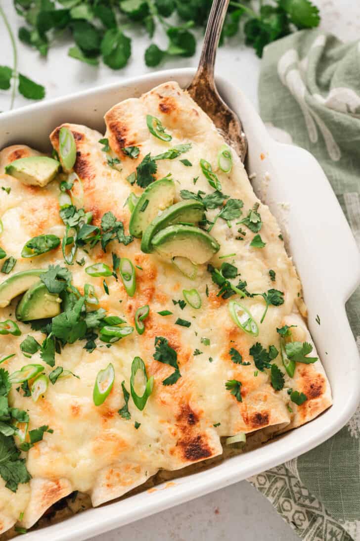 A white baking dish filled with a enchilada recipe vegetarian topped with green herbs and avocado.