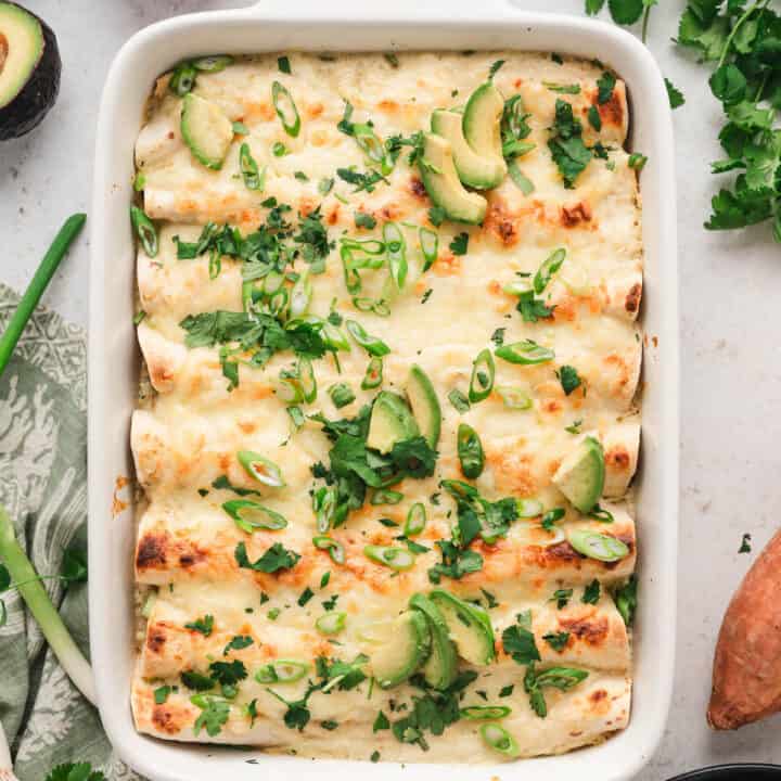 A white baking dish filled with veggie enchiladas topped with green herbs and avocado.