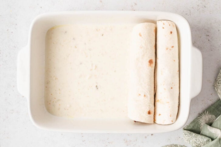 A rectangle white baking dish filled with creamy sauce and two rolled up flour tortillas.