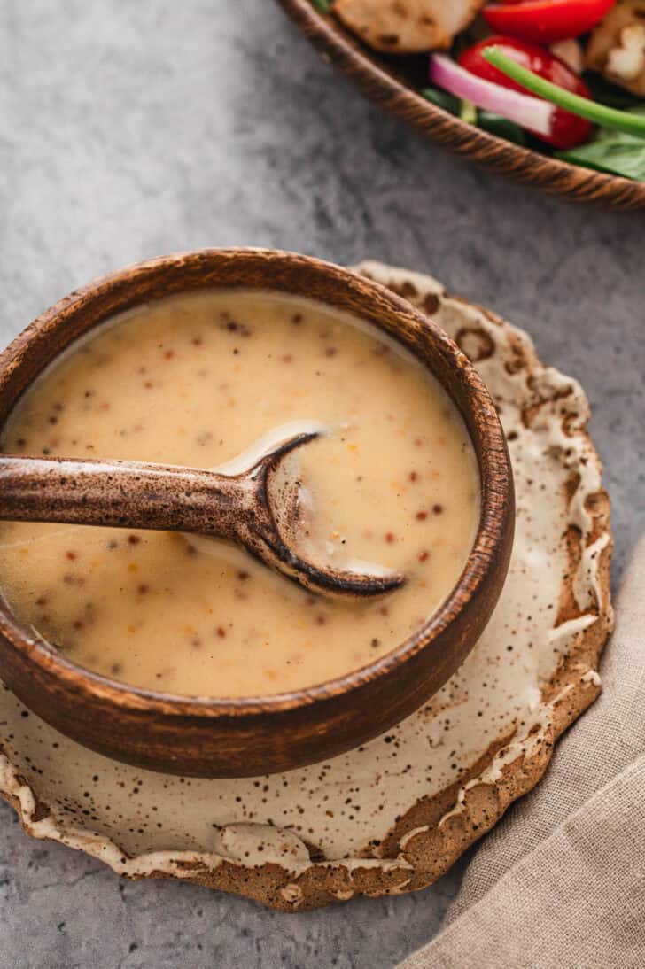 A wooden bowl filled with Dijon honey mustard dressing, with a wooden spoon lifting some out.
