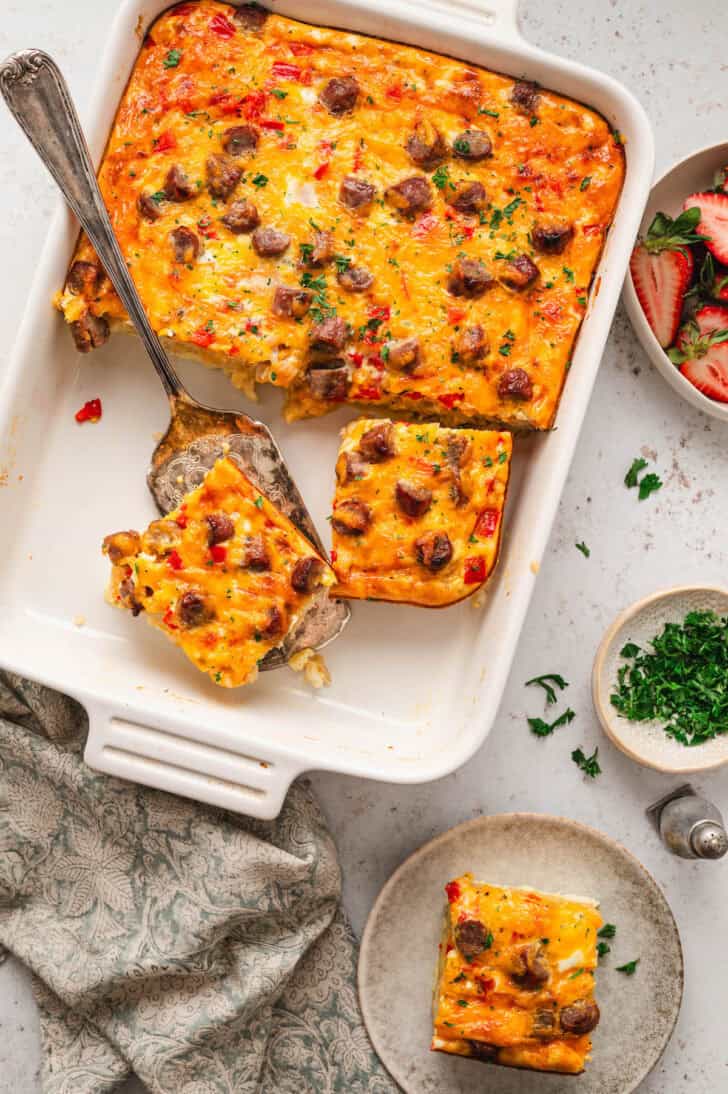 A white rectangular baking dish filled with breakfast casserole with sausage, with a few pieces removed or in the process of being removed.