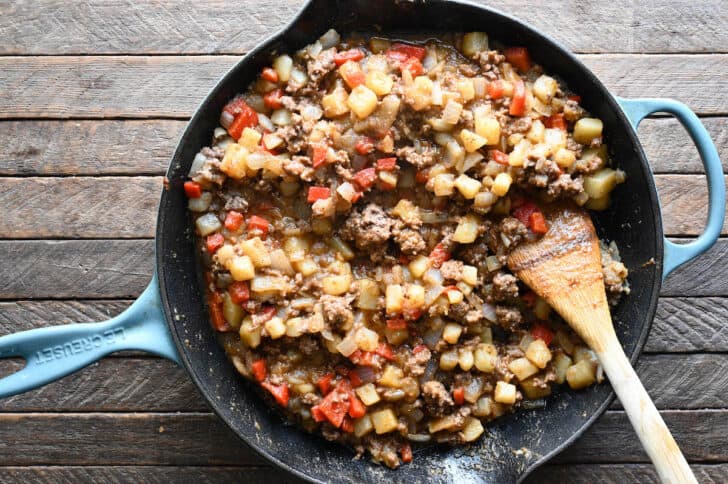 A cast iron skillet with light blue handles, filed with ground beef and potatoes and red bell pepper.