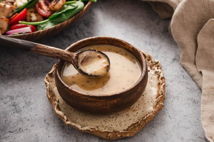 A wooden bowl filled with a honey mustard dressing recipe, with a wooden spoon lifting some out.