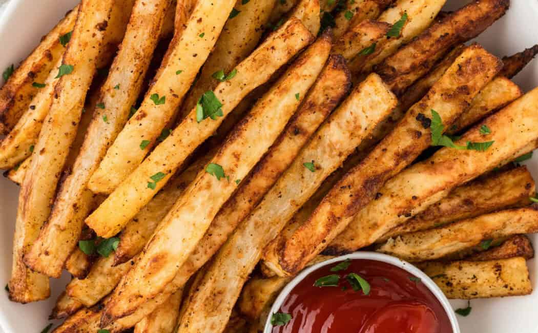 A white plate topped with oven baked French fries with ramekin of ketchup.
